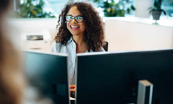 Woman with glasses smiling at desk with computer monitors